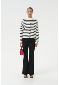 Cardigan a righe bianche e nere con bottoni, abbinato a jeans neri a zampa. La modella tiene in mano una piccola borsa rossa, su uno sfondo semplice.