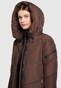 Ruskea toppatakki, jossa on iso huppu, vetoketjusuljin edessä ja mustia metalliyksityiskohtia. Kangas on sileä ja hieman kiiltäväpintainen.