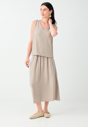 Femme aux cheveux foncés portant un haut beige sans manches à col en V et une longue jupe assortie avec des chaussures plates tissées beige, regardant sur le côté.
