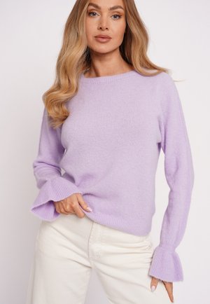 Kobieta o długich, falowanych blond włosach, ubrana w jasnofioletowy sweter z długim rękawem z rozszerzanymi mankietami oraz kremowe spodnie na białym tle.