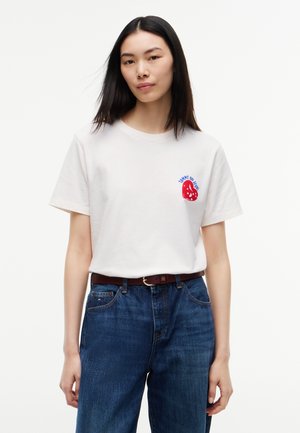 Femme aux cheveux noirs et raides, portant un t-shirt blanc avec un logo rouge et bleu et un jean bleu, debout devant un fond blanc uni.