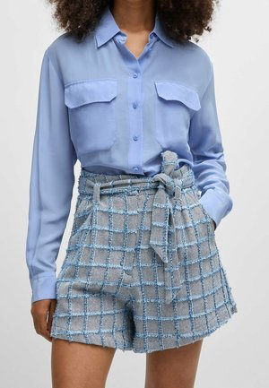 Kobieta ubrana w jasnoniebieską koszulę zapinaną na guziki z klapkowymi kieszeniami, do której dobrano wysokiej talii, teksturowane szorty w kratę, związane w talii.