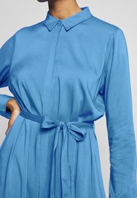 Robe bleue à manches longues avec col, devant boutonné et ceinture nouée à la taille. Le tissu a une texture lisse et brillante avec des plis.