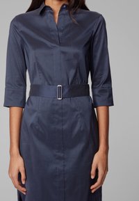 Granatowa sukienka koszulowa wykonana z gładkiego materiału, z kołnierzykiem, rękawami o długości trzy czwarte, wcięciem w talii z srebrną klamrą oraz bocznymi kieszeniami.