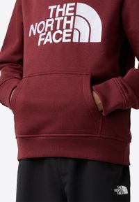 bordová mikina s bielym logom "THE NORTH FACE", predným klokankovým vreckom, rebrovanými manžetami a lemom. Nosí sa s čiernymi nohavicami.