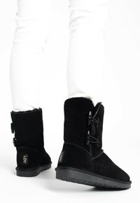 Botas de tobillo de ante negro con un suave forro de piel de piel, que cuentan con dos correas decorativas y una suela de goma texturizada. Logotipo icónico en el tacón.