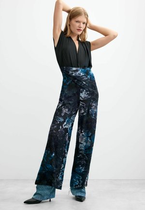 Jeune femme aux cheveux blonds portant un haut noir sans manches et un pantalon large bleu et noir à motifs, posant avec les bras levés derrière la tête.