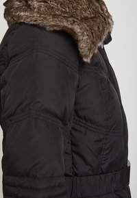 Svart puffjacka med en fuskpälskrage, med belagd midja, quiltad textur och synlig sömdetalj på sidorna.