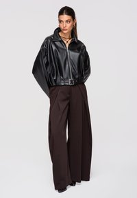 Vrouw draagt een zwarte leren jas met structuur, voorzien van een riem, en een wijde donkerbruine broek, staand tegen een effen lichte achtergrond.