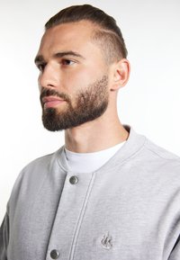 Sweatshirt gri cu un design cu nasturi și o gâtă rotundă. Prezintă o broderie subtilă cu o navă în zona stângă a pieptului. Textură moale.