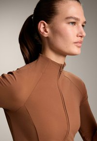 Giacca sportiva marrone con zip e colletto alto, caratterizzata da una texture liscia e un design aderente. Cuciture decorative lungo le spalle e i fianchi.