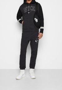 Černá mikina Nike s bílým logem, spárovaná s černými tepláky, které mají logo. Bílá sportovní obuv dokončuje vzhled.