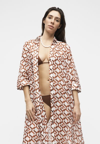 Γυναίκα με μακριά σκουρόχρωμα μαλλιά που φοράει καφέ μπικίνι και λευκό-καφέ πουκάμισο με σχέδιο και ανοιχτά κουμπιά, στέκεται μπροστά σε ένα απλό λευκό φόντο.