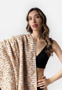 Šal s uzorkom u bež i narančastim tonovima, sa zigzag i točkastim dizajnom. Lagana tkanina elegantno pada preko ramena modela.