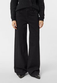 Czarne spodnie o szerokich nogawkach z subtelnym brokatowym zdobieniem, noszone z czarnym dzianinowym swetrem i czarnymi spiczastymi butami.