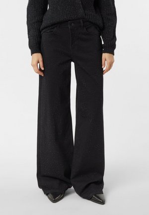 Czarne spodnie o szerokich nogawkach z subtelnym brokatowym zdobieniem, noszone z czarnym dzianinowym swetrem i czarnymi spiczastymi butami.