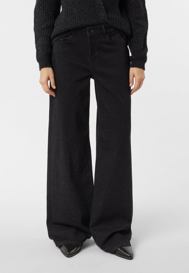 Czarne spodnie o szerokich nogawkach z subtelnym brokatowym zdobieniem, noszone z czarnym dzianinowym swetrem i czarnymi spiczastymi butami.