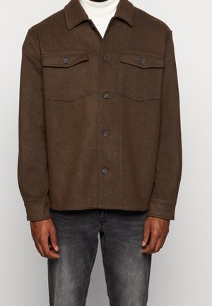 Homme portant une veste marron boutonnée avec deux poches poitrine sur un col roulé blanc, associée à un jean noir délavé.