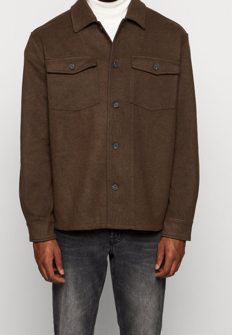 Homme portant une veste marron boutonnée avec deux poches poitrine sur un col roulé blanc, associée à un jean noir délavé.