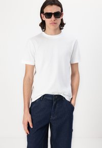 Fehér, rövid ujjú póló kerek nyakkivágással és kis logó részlettel, sötétkék, kényelmes fazonú farmerrel párosítva, amely denim textúrával rendelkezik.