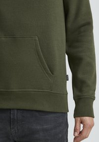 Olive green hoodie with a front pocket, ribbed cuffs, and hem. Made of soft fabric with a smooth texture, featuring a branded tag.