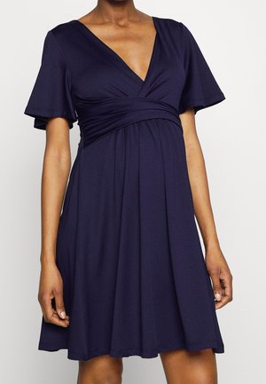 Femme portant une robe bleu marine à manches courtes, col en V, longueur genoux, avec une taille ajustée et une jupe fluide, sur un fond uni.