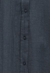 Tumma harmaa kangas, jossa näkyy rakenne, ja neljä mustaa nappia on ommeltu pystysuoraan riviin.