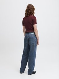Person med bølgete brunt hår iført en kortermet maroon topp, brede blå jeans og svarte sko, stående mot en enkel hvit bakgrunn.