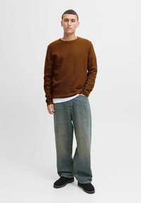 Braun strukturiertes Langarmshirt über einem weißen T-Shirt, kombiniert mit weiten hellblauen Jeans und schwarzen Schuhen. Das Modell steht vor einem schlichten Hintergrund.