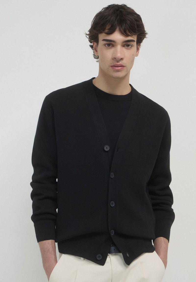 Junger Mann in schwarzem, knöpfbarem Cardigan über schwarzem Hemd und beigen Hosen, steht mit den Händen in den Taschen vor einem schlichten Hintergrund.