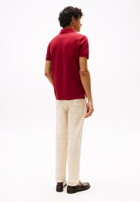 Rdeča polo majica s kratkim ovratnikom, kombinirana s bež chino hlačami. Obleko dopolnjujejo temno rjavi loaferji. Čist dizajn, brez vidnih vzorcev.