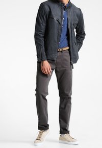 Schwarze Lederjacke mit Reißverschlusstaschen, blau gestreiftes Hemd, graue Hose und cremefarbene Sneakers. Fokus auf einem eleganten Design und einer taillierten Silhouette.