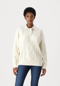 Cremfarbener Strickpullover mit kragenförmigem Halsausschnitt, der ein strukturiertes Muster und diamantförmige Designs auf dem Körper aufweist. Lange Ärmel.