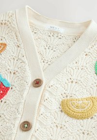 Cárdigan de punto en color crema con un patrón de encaje, que presenta apliques de frutas de colores y dos botones marrones en el frontal.