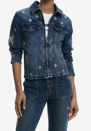 Denimjakke og jeanssæt med broderede blomster- og geometriske mønstre, båret over en hvid skjorte.