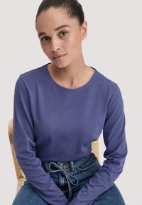 Langärmliges lila Oberteil aus weichem Stoff mit Rundhalsausschnitt, kombiniert mit hoch taillierten Jeans aus Denim mit einer gebundenen Taille.