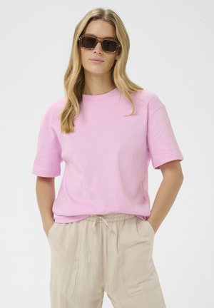 Hosszú szőke hajú nő barna, túlméretezett napszemüveggel, laza, rózsaszín rövid ujjú ingben és bézs színű, zsinórral szabályozható nadrágban, kezei a zsebekben.