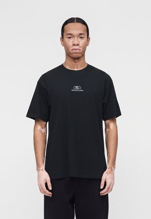 Jaunas vyras su vitiligu, dėvintis juodą „Rip Curl“ marškinėlį, stovintis tiesiai prieš paprastą šviesią foną.