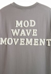 Camiseta de algodón gris con las palabras "MOD", "WAVE" y "MOVEMENT" en letras blancas texturizadas con un diseño en relieve.
