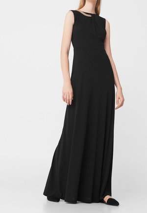 Femme portant une robe noire sans manches longue jusqu'au sol avec un détail de chaîne dorée près du col et des chaussures plates noires.