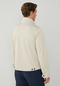 Lys beige jakke med stålende krave, synlig syning, sidelukninger og en glat tekstur. Bagfra ses en skræddersyet pasform.