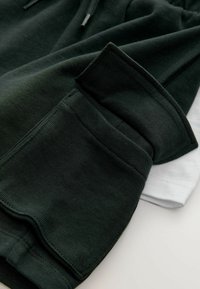 Dos sudaderas dobladas: una verde oscuro con una textura suave y una blanca debajo. Cuenta con puños acanalados y cordones ajustables.