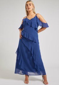 Lång, flödande klänning i djupt blått, tillverkad av chiffon. Har en volangdesign, axelbarar ärmar och en lagerstruktur.