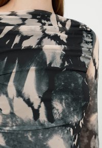 Gros plan d’une personne portant un vêtement en tissu à motif tie-dye noir et gris avec des textures plissées superposées autour de l’épaule et de la poitrine.