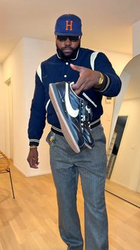 Ténis Nike em couro preto com o swoosh branco e atacadores creme, apresentando uma sola texturizada em borracha castanha. Pessoa segurando os ténis numa sala bem iluminada.