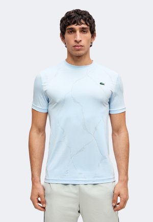 Giovane uomo con capelli scuri e ricci che indossa una camicia azzurro chiaro a maniche corte e pantaloni chiari, in piedi davanti a uno sfondo semplice.