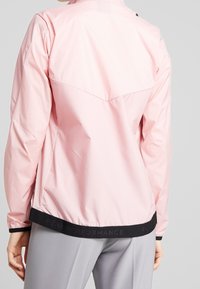 Hellrosa Jacke aus glattem, leichtem Material mit schwarzem Saum. Verfügt über einen hohen Kragen und reflektierendes Branding auf der Rückseite.