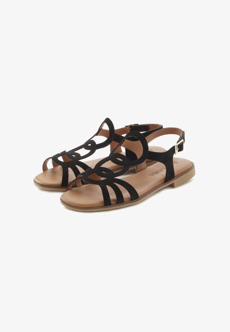 Black suede sandals with geometric cut-out design, flat sole, and tan footbed. Features buckle strap for adjustment and stability.