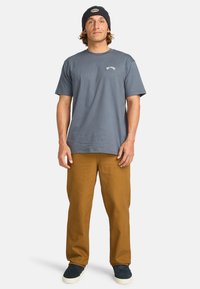 T-shirt grigio in cotone a maniche corte con logo frontale, abbinato a pantaloni marroni e scarpe blu navy. L'individuo indossa un berretto navy.