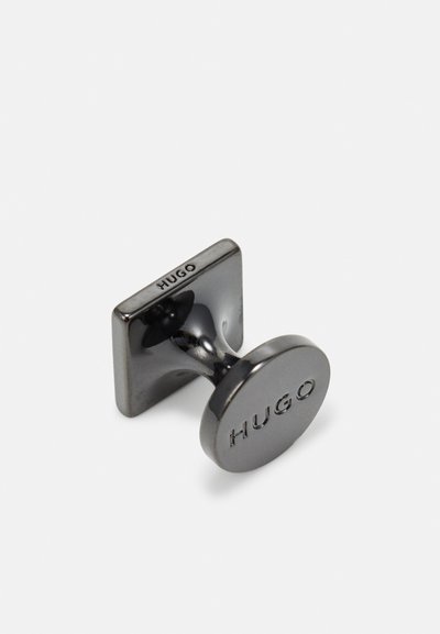 Bouton de manchette en métal noir avec un dos carré et un devant circulaire, embossé avec "HUGO." Surface lisse avec une finition réfléchissante.